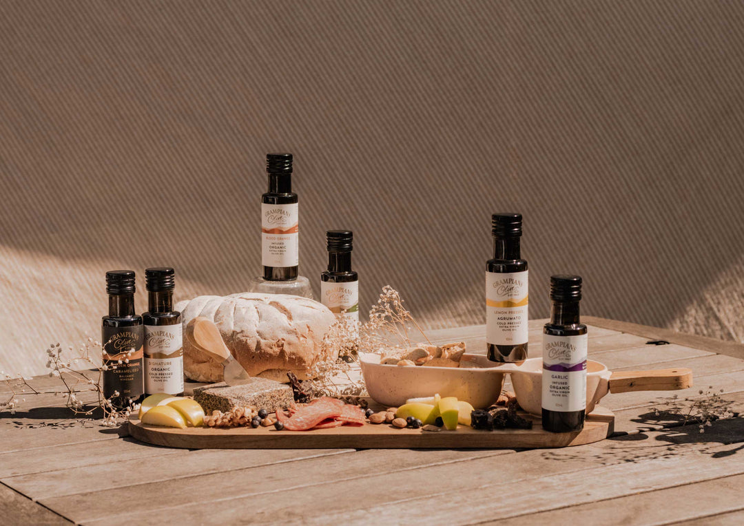 Bottles of olive oil and balsamic vinegar on a wooden tray with bread and fruits against a neutral background