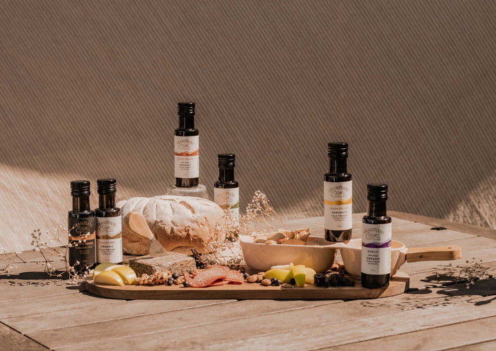 Bottles of olive oil and balsamic vinegar on a wooden tray with bread and fruits against a neutral background